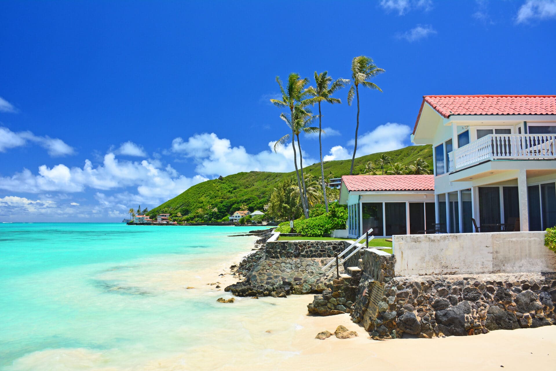 Oceanfront beach home with turquoise waters and swaying palm trees on a clear sunny day at Lanikai Beach on the windward side of Oahu, Hawaii.